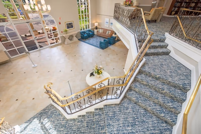 View from the top of a carpeted staircase with ornate gold and black railing leading down to a spacious lobby area with tiled floors, a round table with a flower arrangement, and a seating area with blue chairs and a brown sofa near large arched windows letting in natural light.