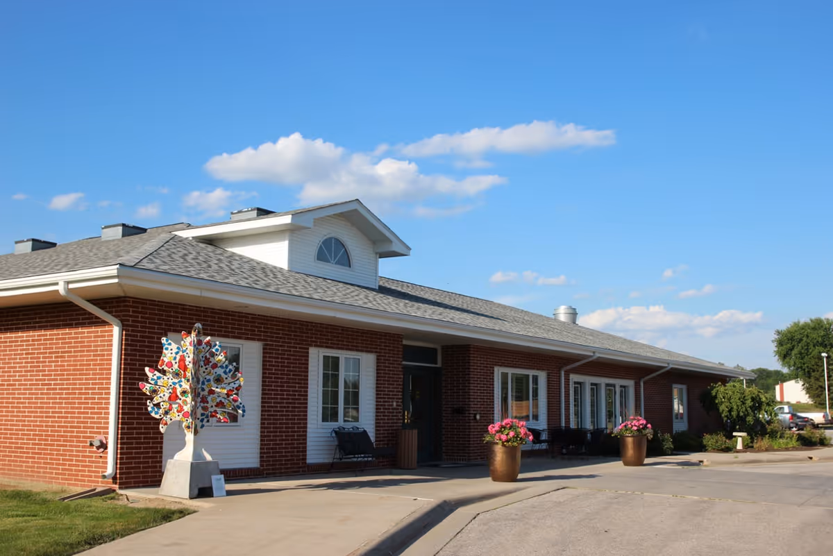 Exterior view of a single-story brick building with a gray shingled roof under a clear blue sky. The building has white-framed windows and a small porch area with benches and large flower pots containing pink flowers. A colorful decorative tree sculpture is placed near the corner of the building.