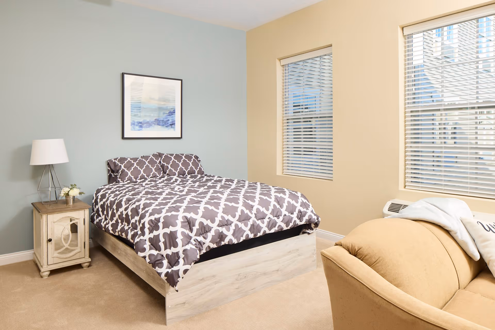 A bedroom with a bed featuring a gray and white patterned comforter and matching pillows. Next to the bed is a small wooden nightstand with a white lamp and a small flower arrangement. The walls are painted in two colors: light blue behind the bed and beige on the adjacent wall with two windows covered by white blinds. A beige armchair is partially visible in the foreground.