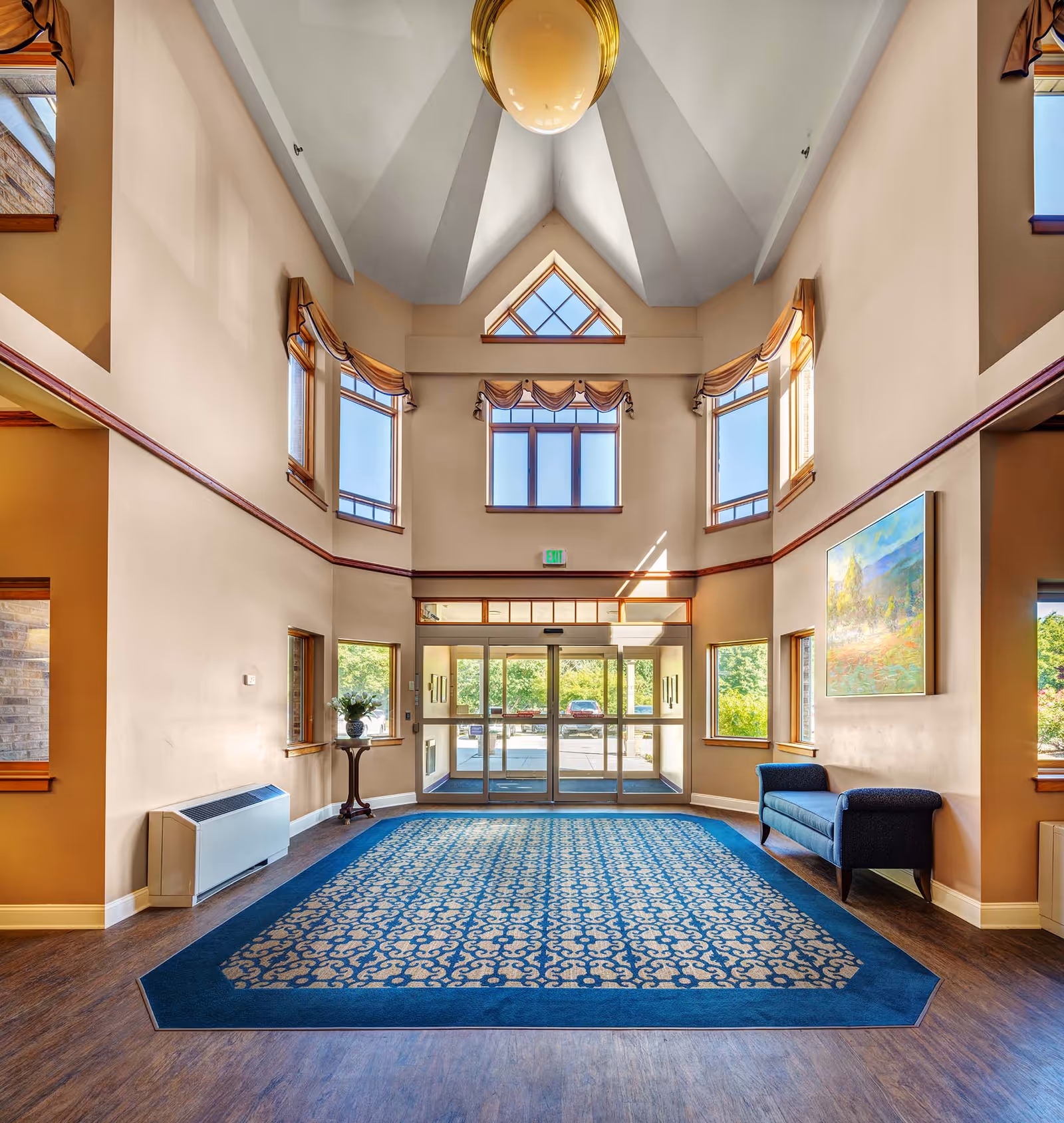 Bright two-story lobby with a large patterned blue rug, glass entrance doors, vaulted ceiling, seating bench and wall art.