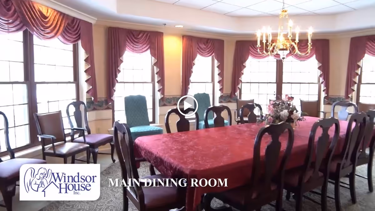 A large dining room with a long table covered by a red tablecloth surrounded by wooden chairs. The room has multiple large windows with red drapes and valances, a chandelier hanging from the ceiling, and a floral centerpiece on the table.