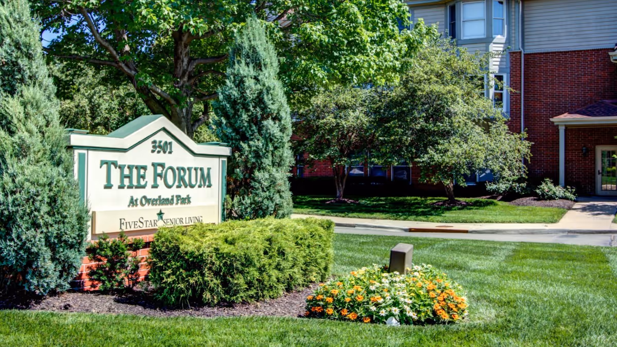 Outdoor view of The Forum at Overland Park senior living facility sign surrounded by green bushes, colorful flowers, and trees with part of the building visible in the background.