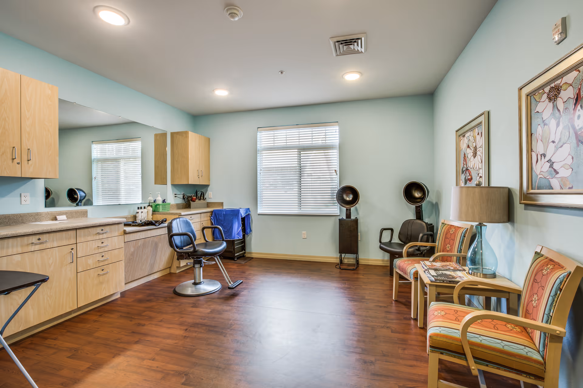 Bright interior salon area with styling chairs, countertop and cabinets, hooded hair dryers, and a small seating area with armchairs and a lamp.