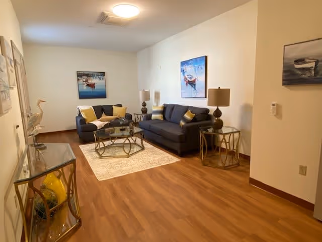 A cozy living room with two dark blue sofas adorned with yellow and white pillows, a glass coffee table on a light-colored rug, two side tables with matching lamps, wooden flooring, and framed artwork of boats on the walls.