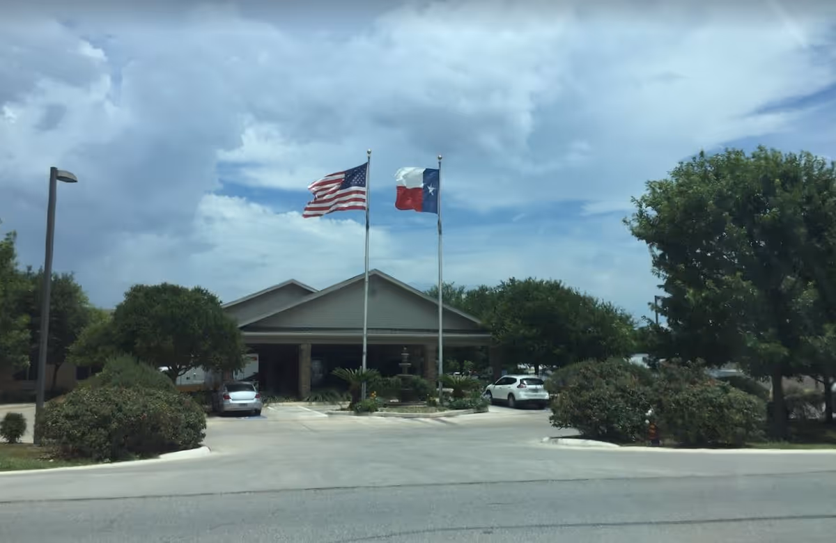 Front exterior view of Silver Tree Nursing and Rehabilitation facility with two flagpoles displaying the American and Texas flags, surrounded by trees and bushes, with a driveway and parked cars in front.