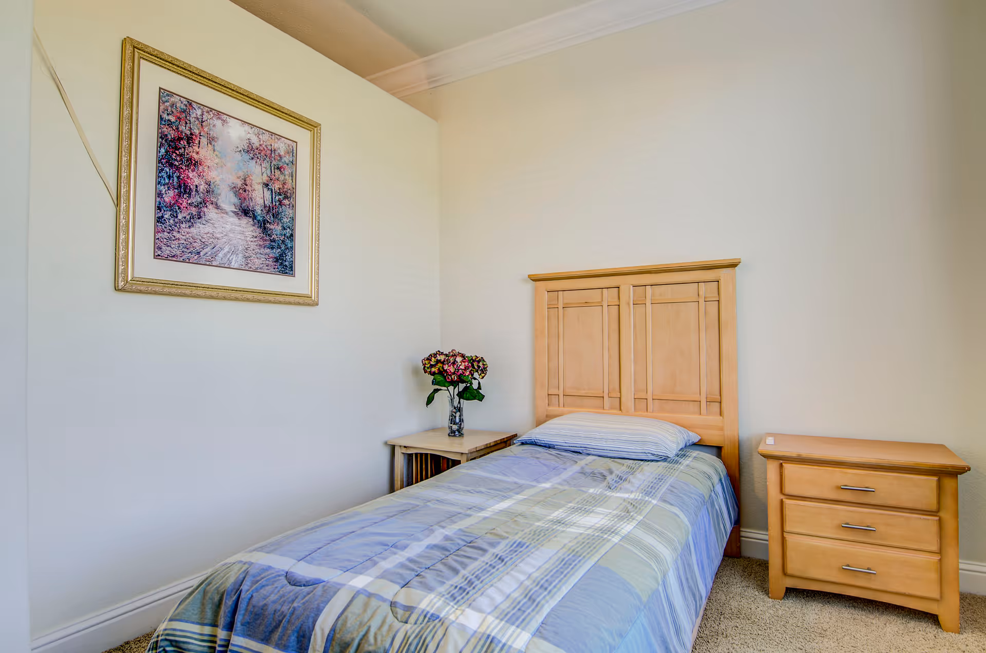 A simple bedroom with a single bed featuring a wooden headboard and a blue and green plaid comforter. There is a wooden nightstand with three drawers on the right side of the bed and a small table with a vase of flowers on the left. A framed painting of a forest path with colorful trees hangs on the wall above the left side of the bed.