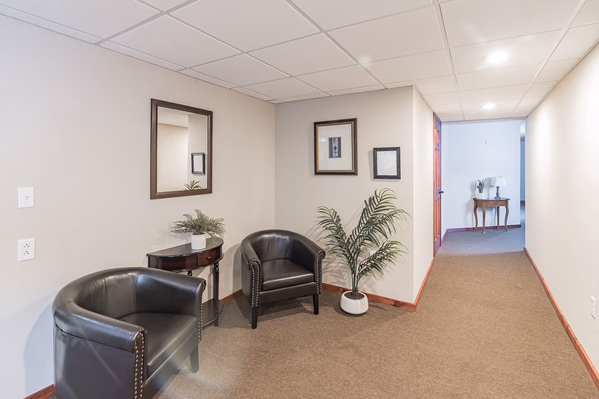 A hallway area in Briarwood Senior Living with beige walls and carpeted floor. Two black leather armchairs are placed near a small dark wooden table with a potted plant. A mirror and two framed pictures hang on the wall above the chairs. Further down the hallway, there is a small wooden table with a lamp and another potted plant.