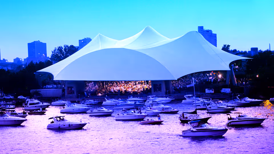 Many small boats anchored on a river in front of a large white tensile-roof concert pavilion at dusk.