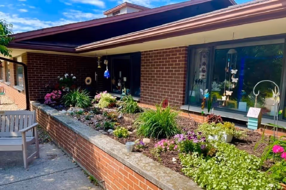 Outdoor garden area with a variety of flowering plants and greenery in a raised brick planter bed next to a brick building with windows. A wooden bench is placed on the concrete walkway beside the garden. The sky is blue with some clouds.