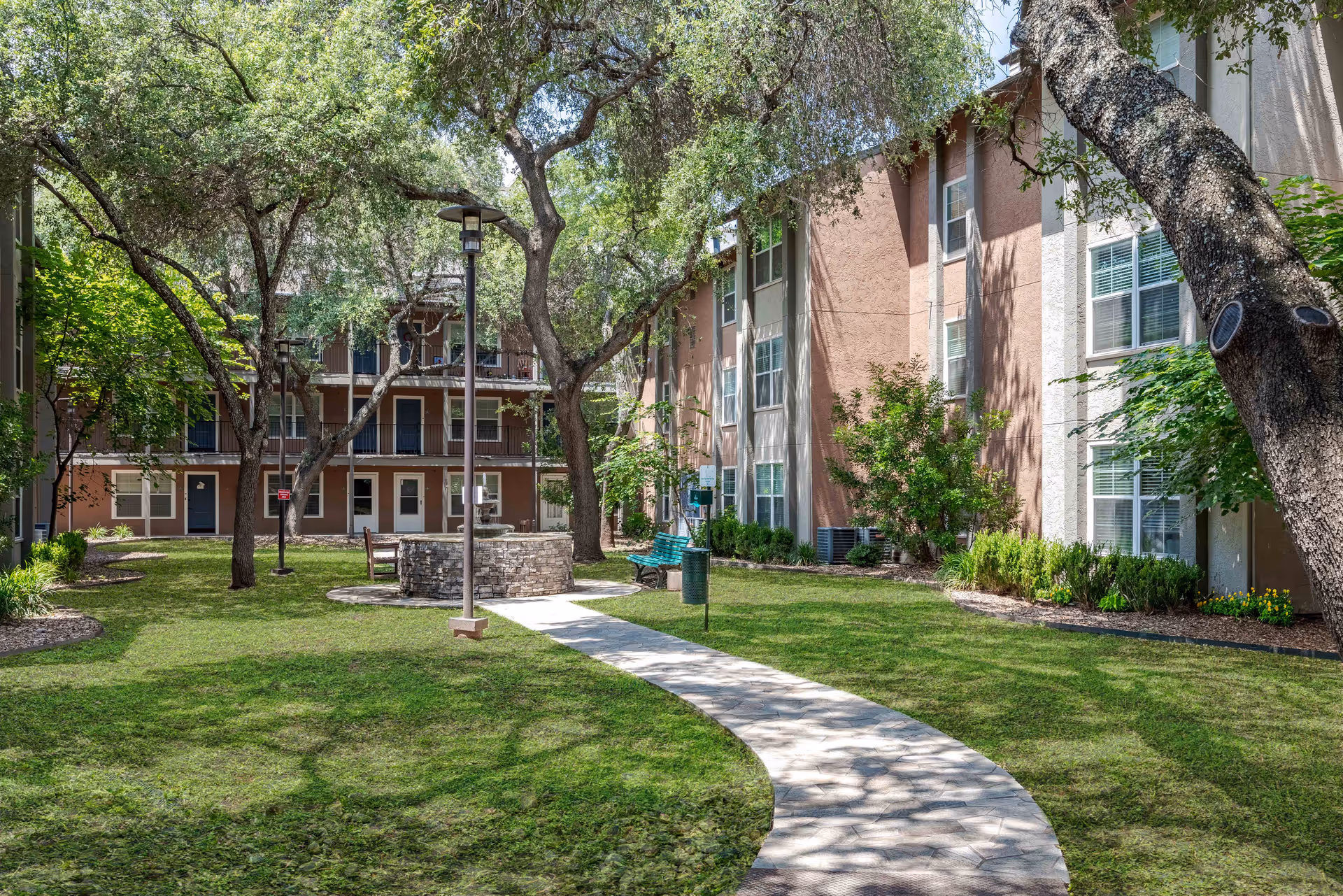 A landscaped courtyard with a curved paved path, grassy lawn, mature trees, benches and seating surrounded by a multi-story residential building.