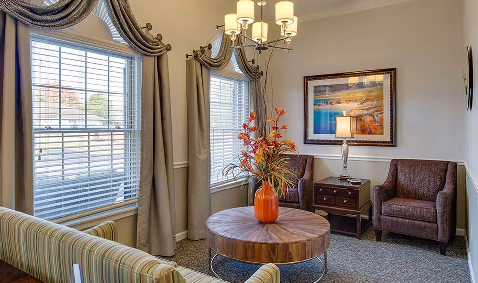 A cozy sitting area with two brown armchairs, a round wooden coffee table with an orange vase holding autumn-colored flowers, a side table with a lamp, and two large windows with beige curtains and blinds. A framed landscape painting hangs on the wall.