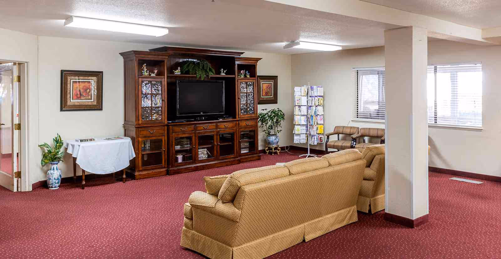 Spacious senior living common lounge with upholstered sofas facing a large wooden entertainment cabinet and TV.