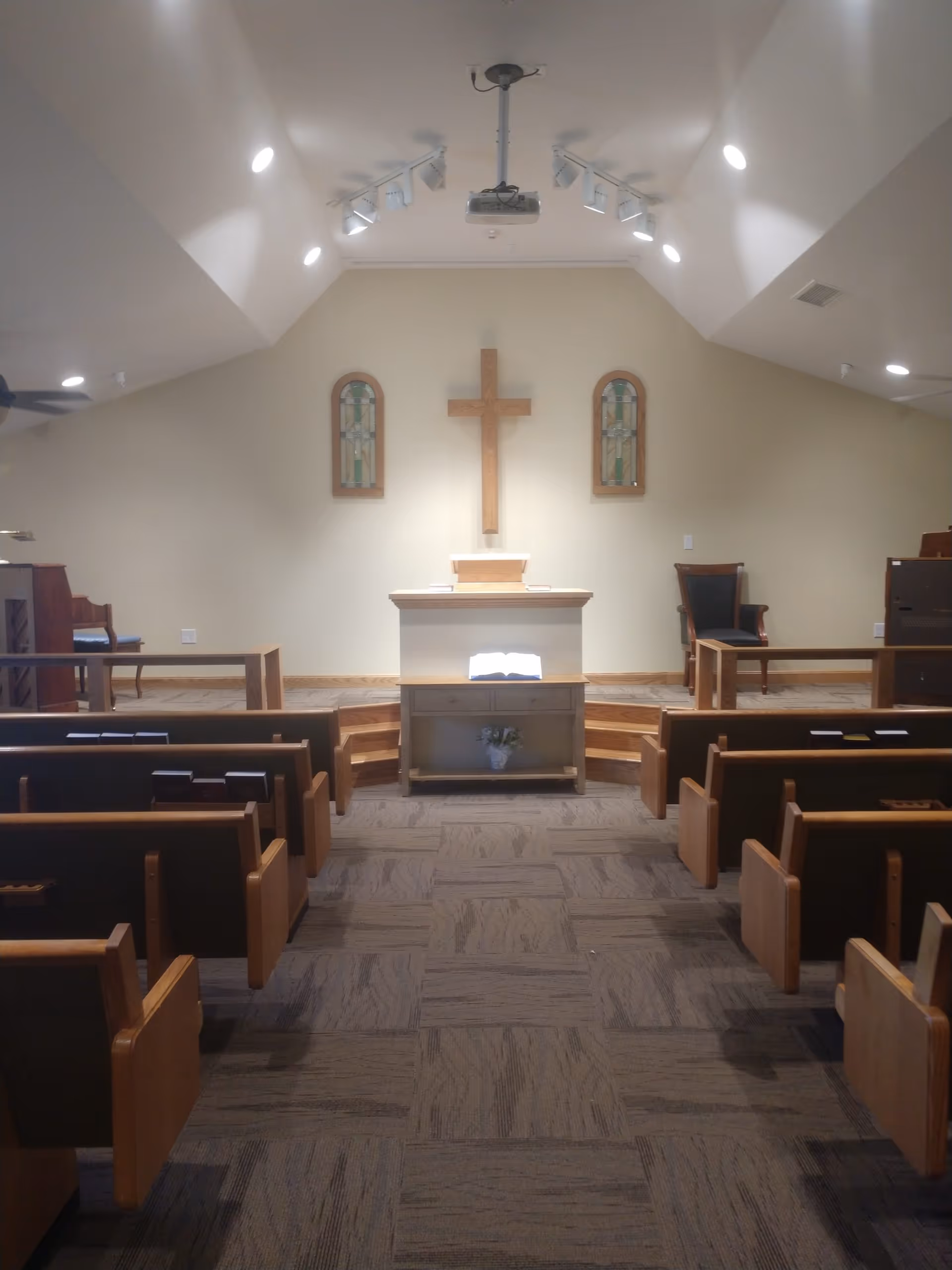 Small chapel interior with wooden pews facing an altar topped by a wooden cross and flanked by stained-glass windows.