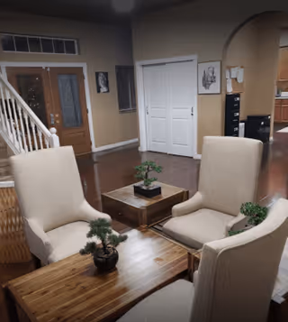 A cozy living room area with four beige upholstered chairs arranged around two wooden coffee tables. Small potted plants are placed on the tables. In the background, there is a wooden front door with glass panels, a staircase with white railings, and a white double-door closet. The walls are painted beige, and there are framed pictures hanging on them. The floor is a polished dark wood.