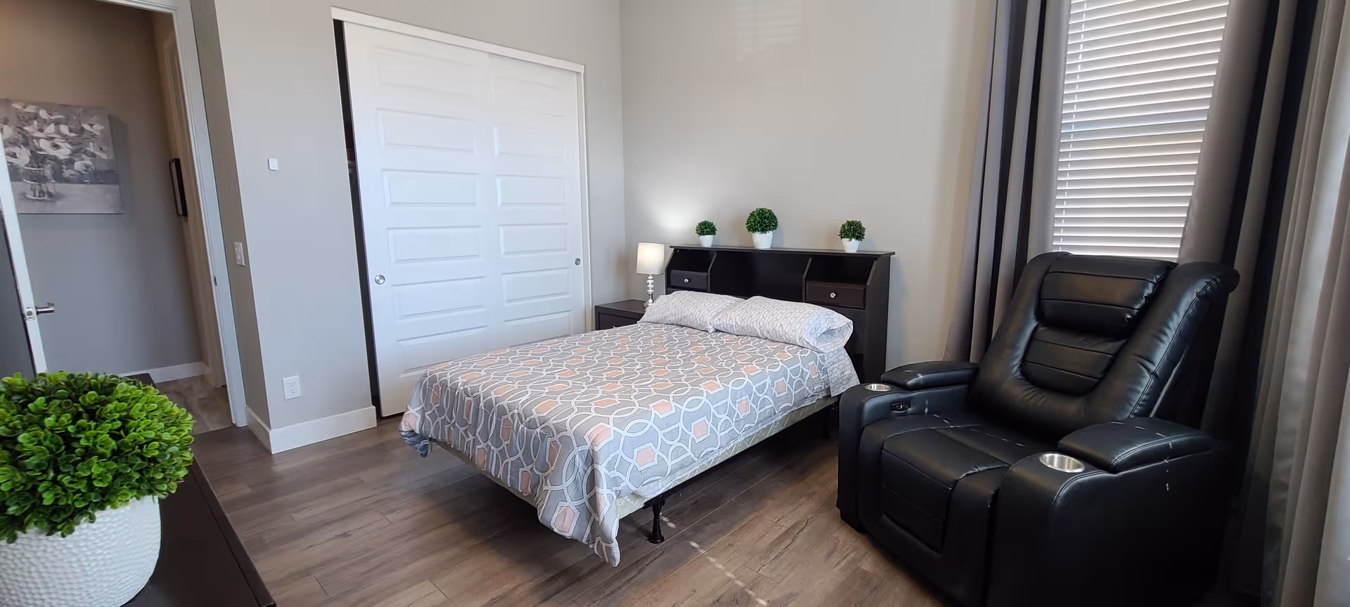 A bedroom with a bed covered in a patterned gray and white bedspread, a black headboard with three small potted plants, a bedside lamp, a black recliner chair with cup holders, a window with blinds and curtains, and a white double-door closet.