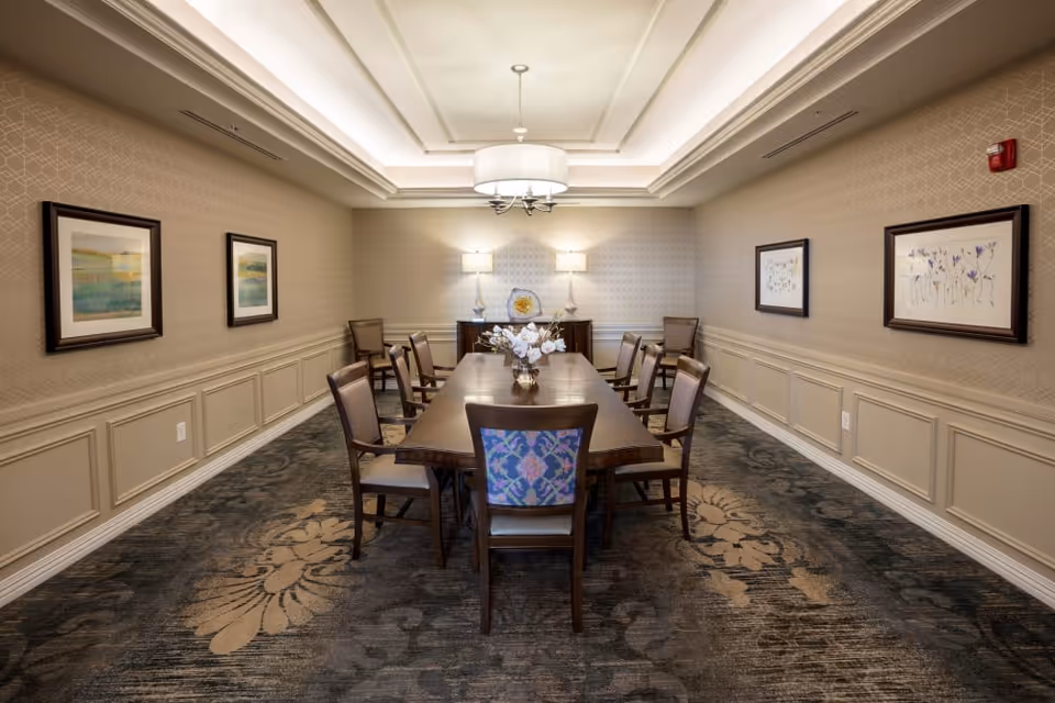 A formal dining room with a long wooden table surrounded by ten chairs, two table lamps on a sideboard at the far end, framed artwork on the walls, and a decorative ceiling light fixture.