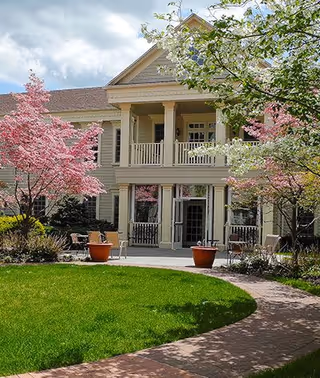 Front of a two-story residential building with balconies overlooking a circular lawn, pink flowering trees, potted plants, and outdoor seating.