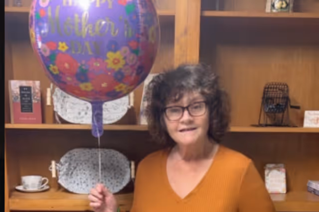 An elderly woman with curly hair and glasses wearing an orange sweater is holding a purple balloon that says 'Happy Mother's Day' with colorful flowers printed on it. She is standing in front of a wooden shelf with decorative plates, a teacup, and other small items.