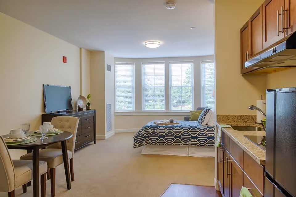 A bright studio apartment with a small dining table set for two, a dresser with a TV on top, a bed with patterned bedding near large windows, and a kitchenette with wooden cabinets and a small refrigerator.