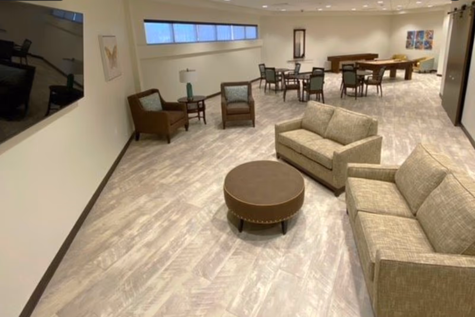 A spacious common area with beige sofas, brown armchairs, a round brown ottoman, several tables with chairs, a lamp on a side table, a large wall-mounted TV, and a pool table in the background. The room has light-colored flooring and white walls with some artwork.