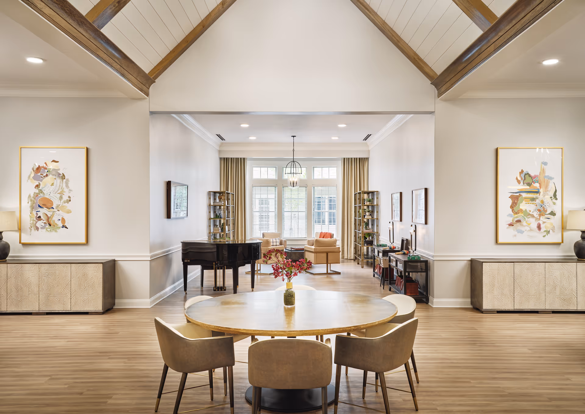 Bright communal living room and dining area with a round table and chairs in the foreground, a grand piano and seating area under vaulted ceilings with exposed wooden beams.