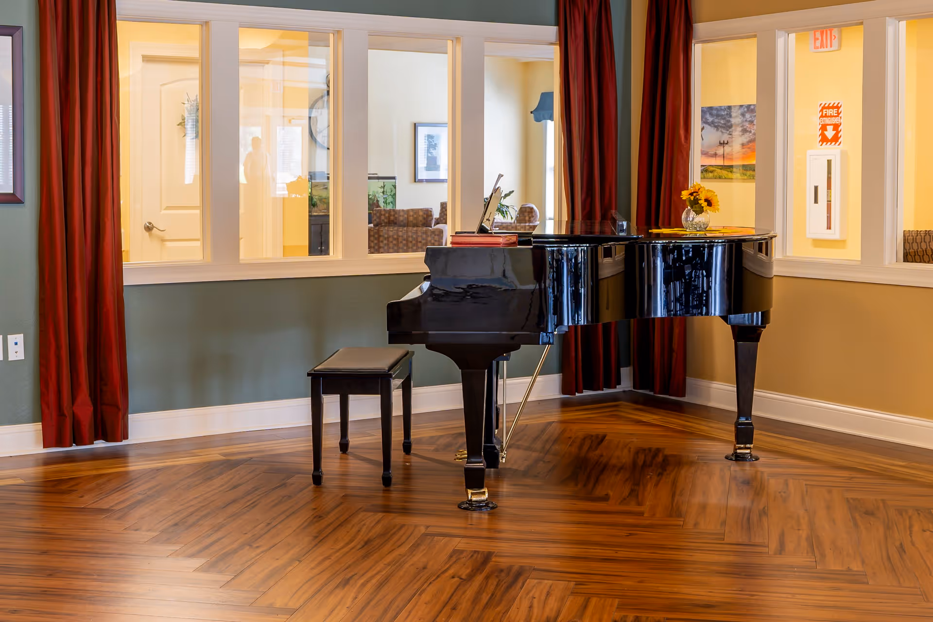 A black grand piano with a matching bench sits on a polished wooden floor in a room with green and beige walls. The room has large windows with white frames and red curtains, through which another room with chairs and framed pictures is visible. A small vase with yellow flowers is placed on the piano.