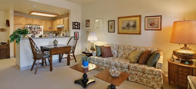 A cozy living room and kitchen area in a senior living facility. The living room features a floral patterned sofa with multiple cushions, two wooden side tables with decorative items, a table lamp on a wooden side table, and framed artwork on the wall. The kitchen area has wooden cabinets, a refrigerator covered with photos and magnets, a stove, and a small dining table with two wooden chairs.