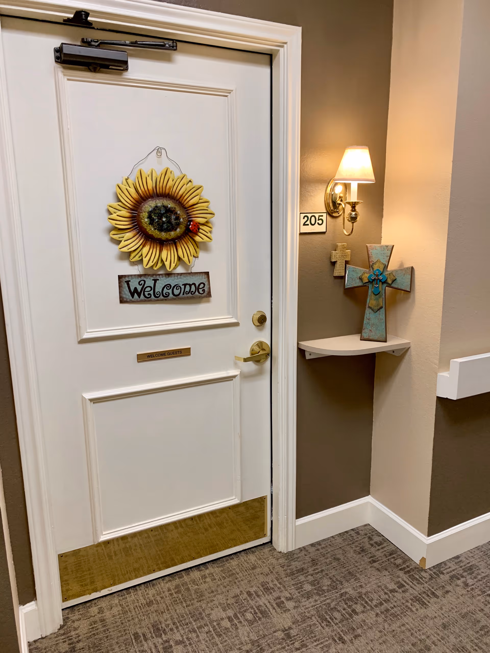A white door with a sunflower decoration and a 'Welcome' sign hanging on it. The door has a brass handle and a small plaque that reads 'WELCOME GUESTS'. To the right of the door is a wall with a room number sign '205', a wall-mounted lamp with a white shade, and two decorative crosses on a small white corner shelf. The walls are painted in beige and brown tones, and the floor is carpeted.