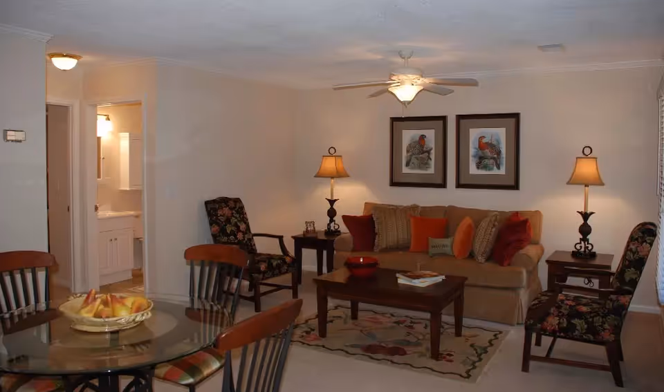 A cozy living room with a beige sofa adorned with multiple cushions, two floral-patterned armchairs, two side tables each with a lamp, and framed bird artwork on the wall. A wooden coffee table sits on a floral rug in front of the sofa. To the left, a glass dining table with wooden chairs is visible, and an open doorway leads to a bathroom.