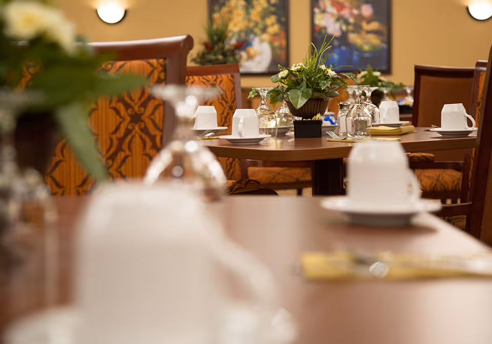 A dining room with wooden tables set with white cups, saucers, upside-down wine glasses, and silverware. The chairs have patterned orange upholstery, and there are decorative plants on the tables. The background features framed artwork on beige walls.