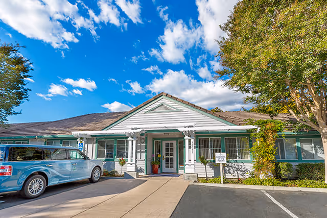 A single-story white senior living building with a centered front entrance and a parked blue SUV.