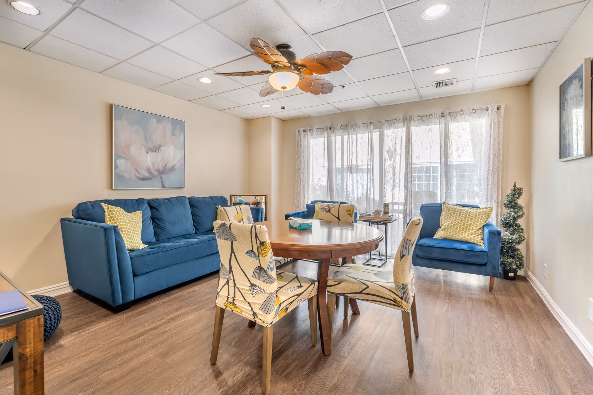 A bright living room with a round wooden table surrounded by four floral-patterned chairs. There are two blue upholstered sofas with yellow patterned throw pillows. A ceiling fan with leaf-shaped blades hangs from the white tiled ceiling. Sheer curtains cover large windows letting in natural light. The room has light-colored walls and wood flooring, with a small decorative plant in the corner and framed artwork on the walls.