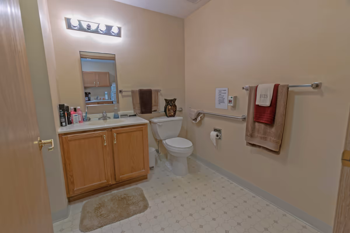A clean bathroom with beige walls and hexagonal tiled floor. There is a wooden vanity with a sink, a mirror above it, and a light fixture with four bulbs. On the countertop are various toiletries. Next to the vanity is a white toilet with a decorative owl figurine on top. Two towel racks are mounted on the walls, each holding folded towels. A grab bar is installed near the toilet, along with a small sign and an emergency pull cord.