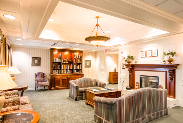 A cozy living room area in a senior living facility with two striped sofas facing a wooden coffee table, a fireplace with decorative items on the mantel, a wooden bookshelf filled with books, and a couple of upholstered chairs. The room is warmly lit with ceiling lights and lamps, featuring neutral-colored walls and carpeted flooring.
