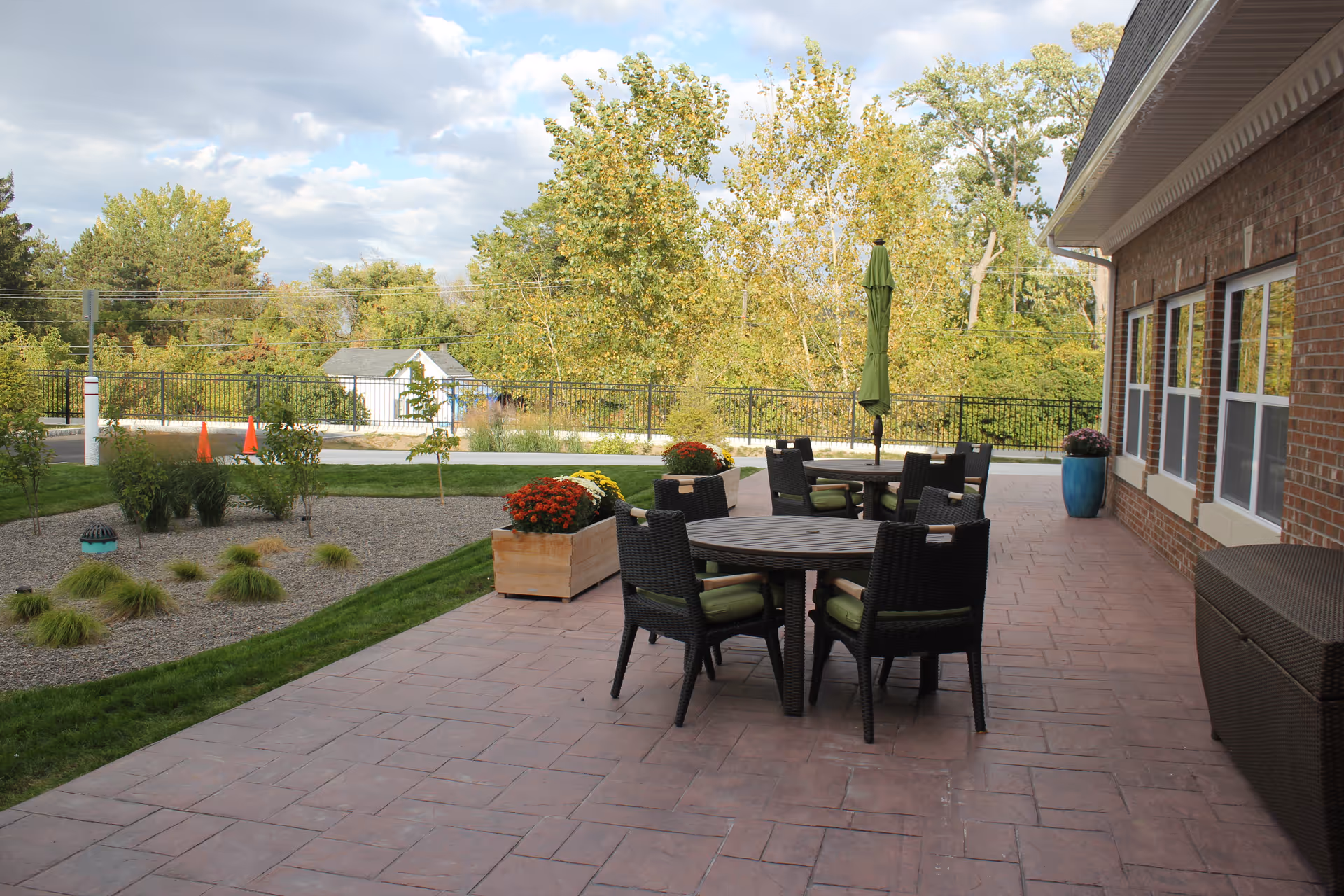 Outdoor patio with round tables and wicker chairs beside a brick building and landscaped yard with trees in the background.