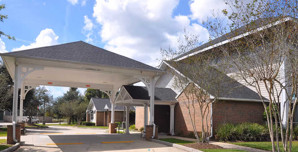 Entrance area of Oak Park Village at Slidell featuring a covered drop-off with white wooden beams and a shingled roof, adjacent to a brick building with small trees and landscaping under a partly cloudy sky.