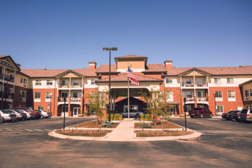 Front exterior view of Lincoln Meadows Senior Living facility showing a three-story building with balconies, an American flag in the center, a covered entrance, and a parking lot with several cars under a clear blue sky.