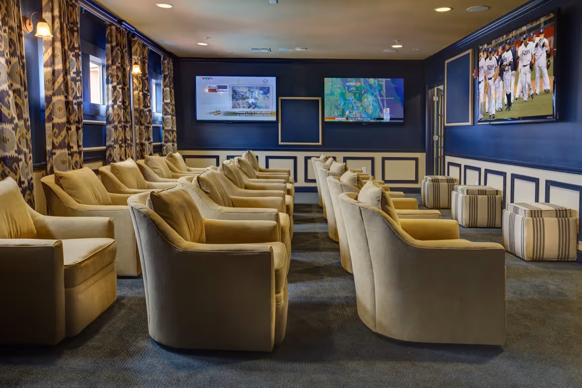 A cozy media room with two rows of beige armchairs facing three wall-mounted flat-screen TVs. The walls are painted dark blue with white paneling, and patterned curtains cover the windows on the left side. There are also several striped ottomans near the front of the room.