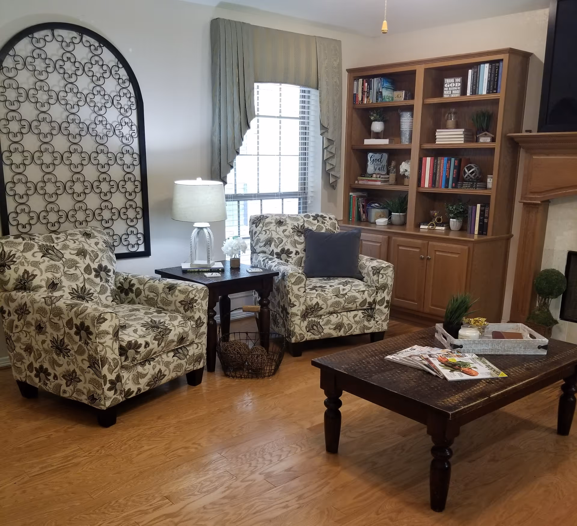 A cozy living room with two floral patterned armchairs, a wooden coffee table with magazines and decorative items, a side table with a lamp and flowers, a large window with curtains, a wooden bookshelf filled with books and plants, and a fireplace with a TV mounted above it.