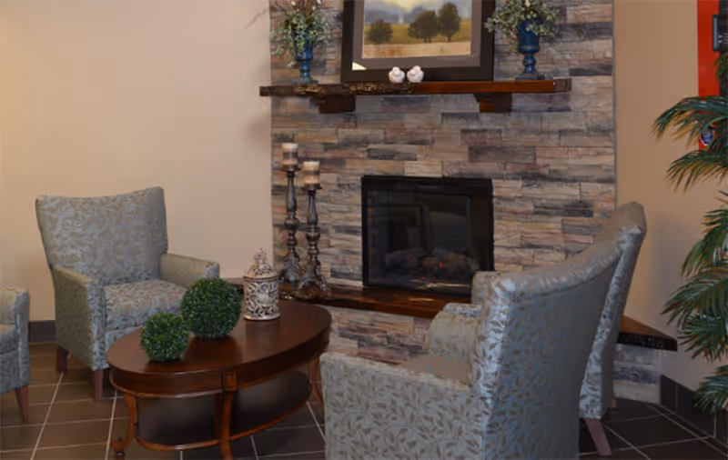 A cozy seating area with three patterned armchairs arranged around a wooden coffee table. The table has decorative green plants and a lantern on it. Behind the chairs is a stone fireplace with a wooden mantel, decorated with candles, small bird figurines, and a framed landscape painting. A potted plant is visible to the right.