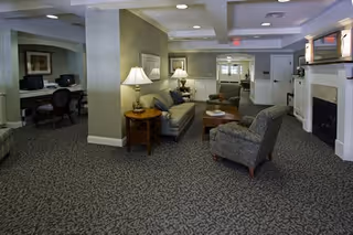 A spacious senior living common area with patterned carpet, a gray armchair, a beige sofa with cushions, two side tables with lamps, a fireplace, and a computer workstation area in the background.
