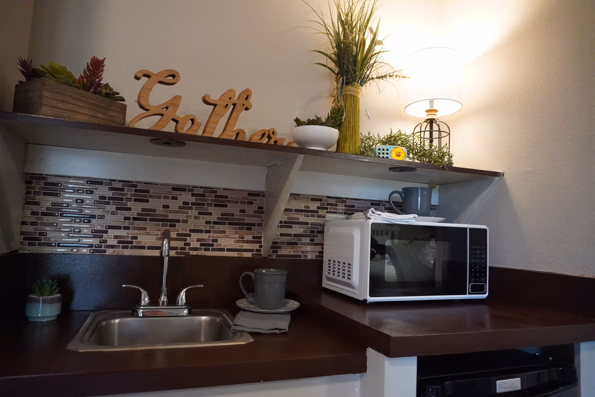 A small kitchenette area with a stainless steel sink, a white microwave on a brown countertop, a gray mug on a saucer with a napkin, and decorative plants on a shelf above. A lamp with a white shade is also on the shelf, casting warm light on the wall.