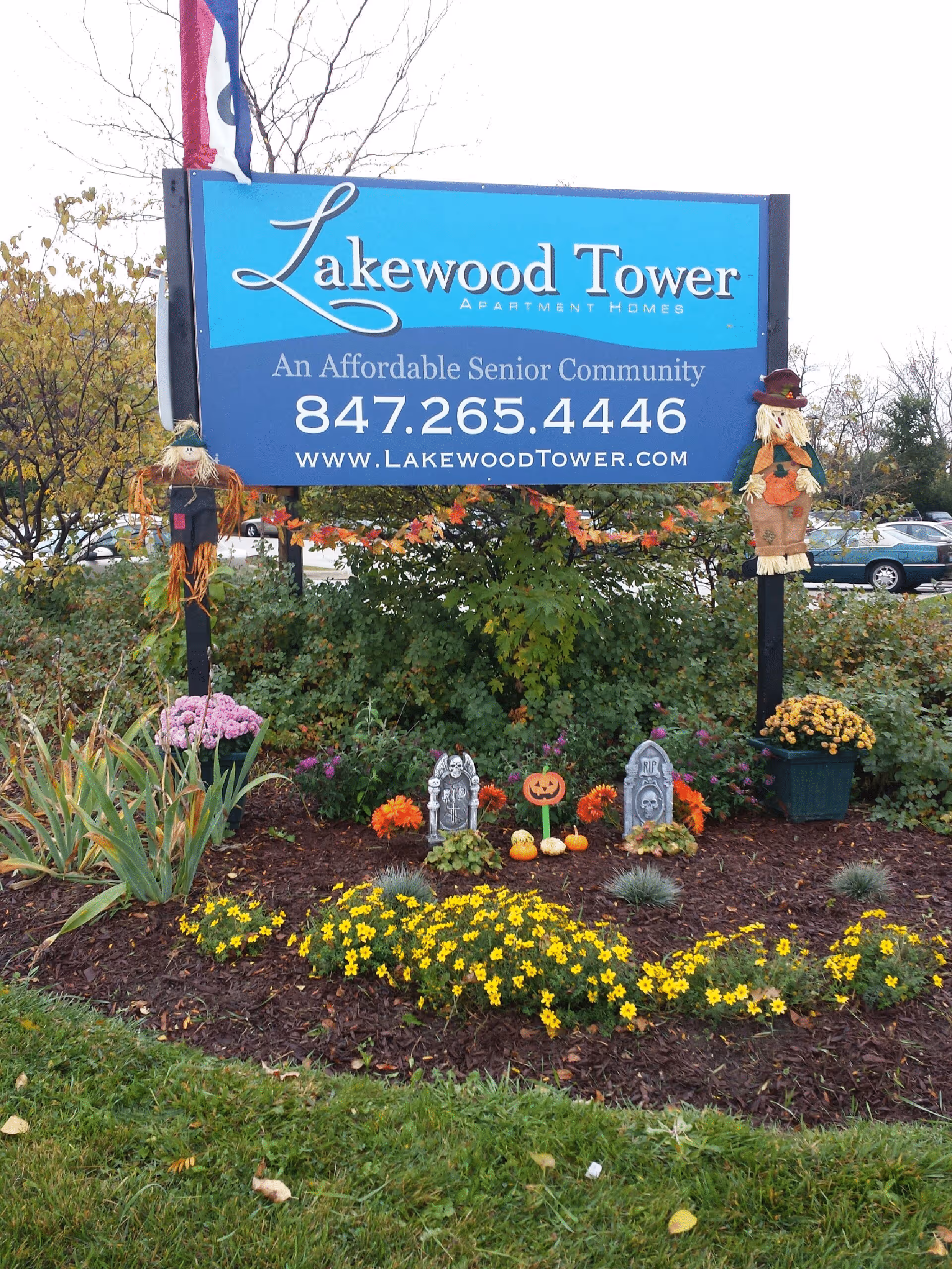 Outdoor sign for Lakewood Tower Apartment Homes, an affordable senior community, with contact phone number and website. The sign is surrounded by fall decorations including scarecrows, flowers, and Halloween-themed items like tombstones and pumpkins.