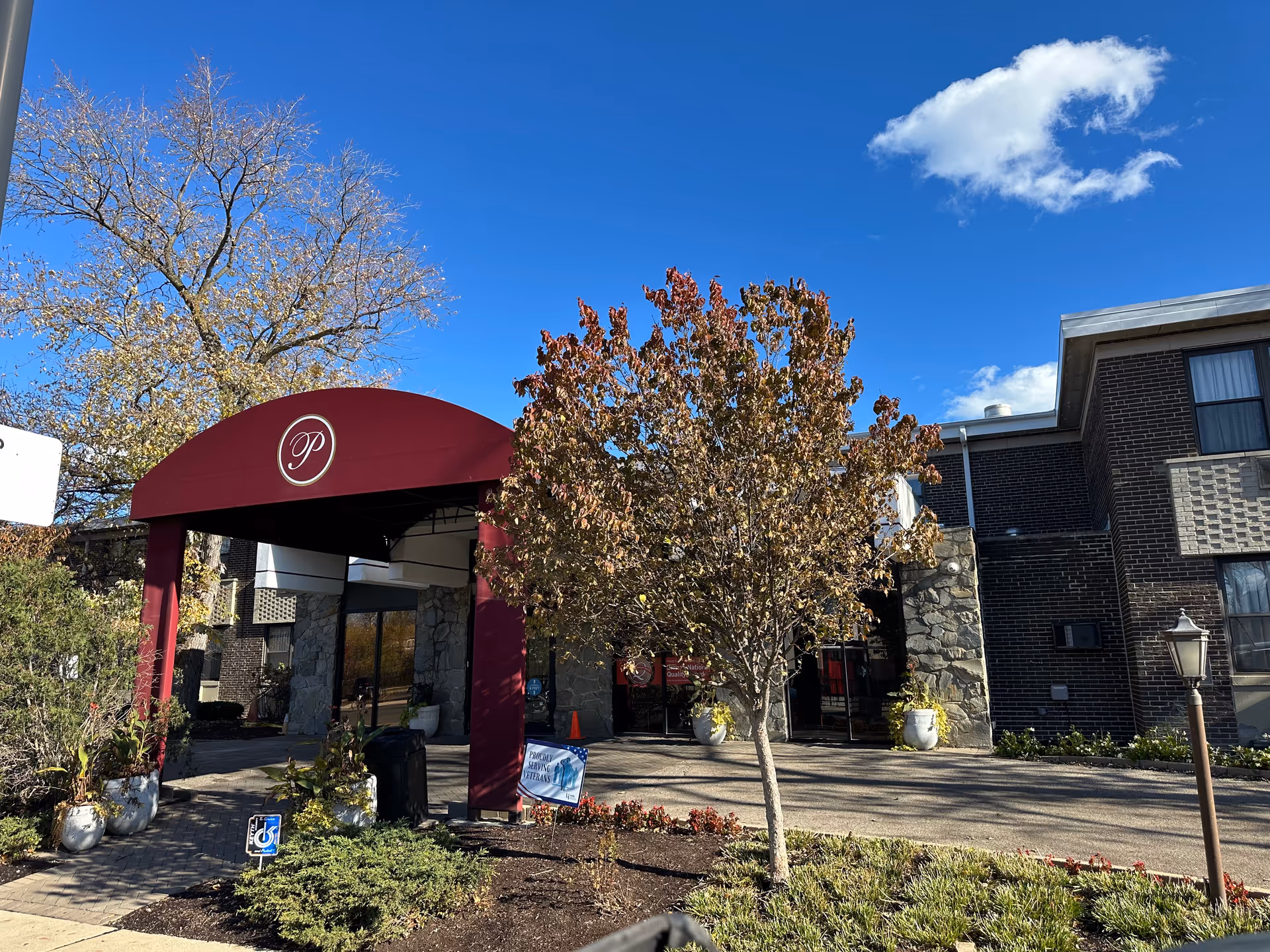 Entrance of Peterson Park Health Care Center with a maroon canopy featuring a white letter 'P'. The building is made of dark brick and stone, with a tree and some plants in front. The sky is clear and blue with a few clouds.