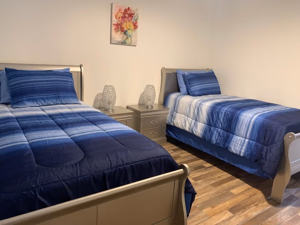 A bedroom with two twin beds featuring blue striped bedding and matching pillows. Between the beds are two gray nightstands, each with a decorative lamp. A colorful floral painting hangs on the white wall above the nightstands. The floor is wooden.