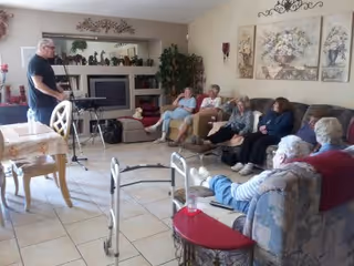 A group of elderly people sitting on couches and chairs in a living room, attentively listening to a man standing near a keyboard. The room has tiled floors, a fireplace with decorative items on the mantle, and framed floral artwork on the wall.