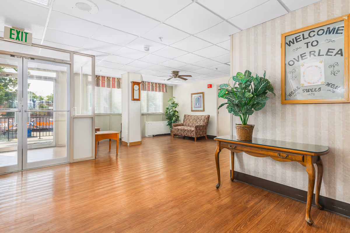 A bright and clean interior common area with wooden flooring, a glass exit door, a wooden table with a potted plant, a patterned armchair, wall clock, ceiling fan, and a framed sign that reads 'Welcome to Overlea Home Sweet Home'.