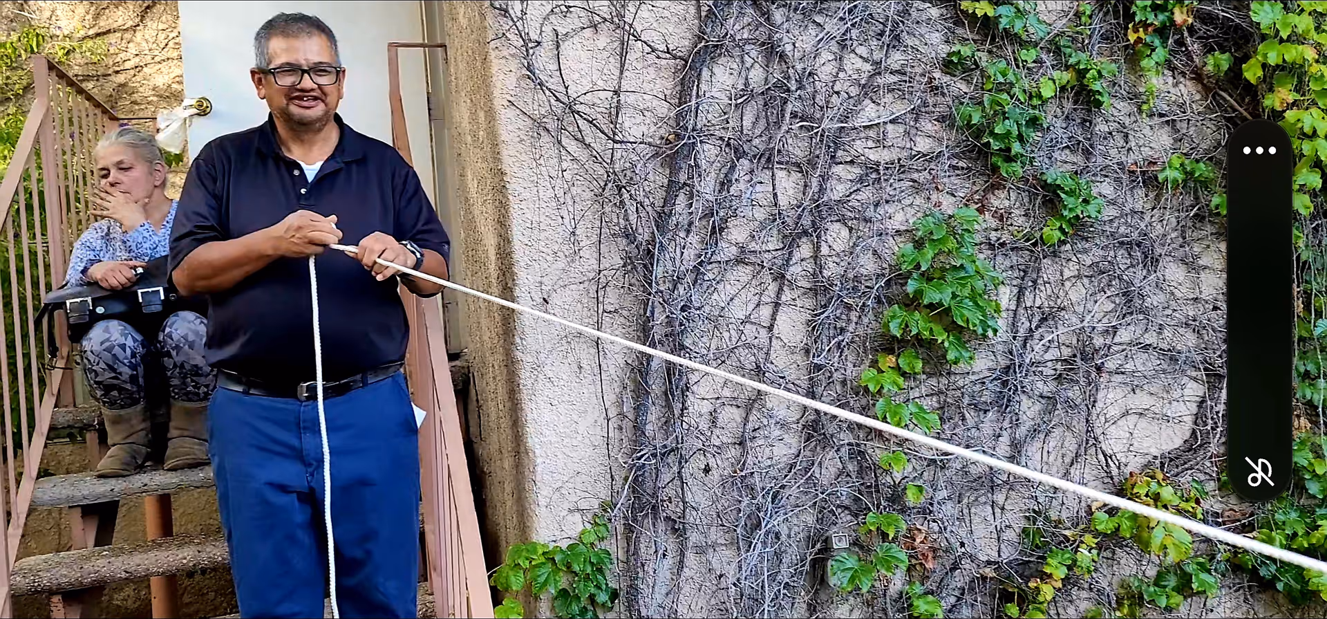 An older man wearing glasses and a black shirt is holding a long white rope outside near a wall covered with dry vines and some green leaves. Behind him, a woman with gray hair is sitting on outdoor stairs, resting her head on her hand.