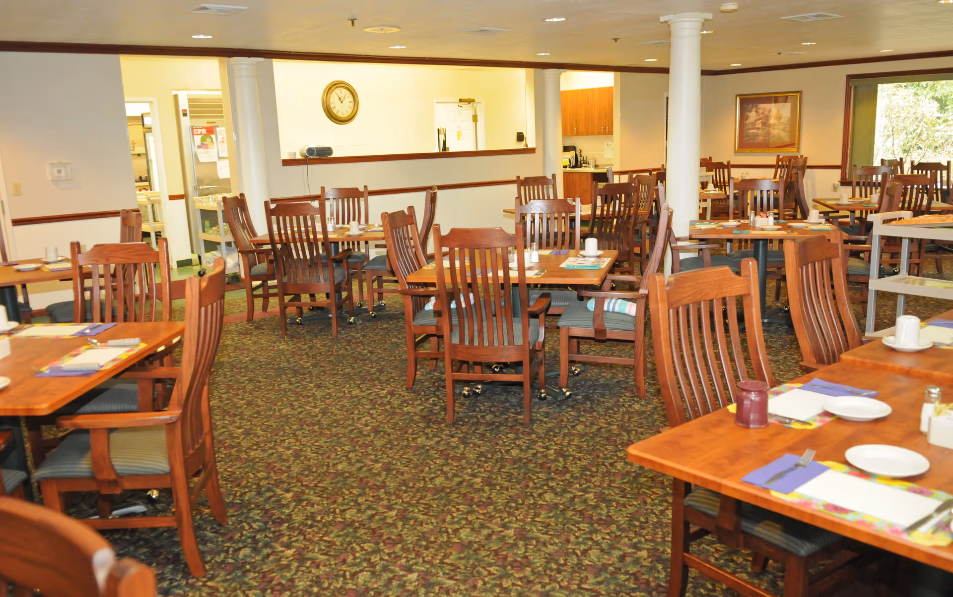 A dining room with multiple wooden tables and chairs arranged neatly. Each table is set with placemats, plates, cups, and utensils. The room has patterned carpet flooring, white walls with wooden trim, and a clock on the wall. There are large windows letting in natural light and a painting on one wall.