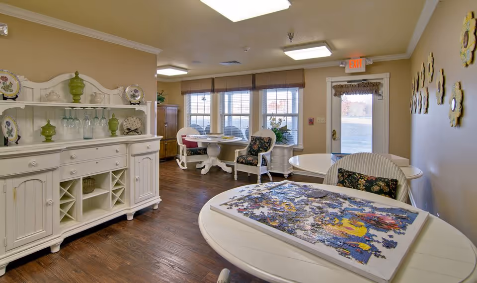A cozy interior room with wooden flooring featuring a white cabinet with decorative plates and green vases, several round tables with floral cushioned chairs, and a partially completed jigsaw puzzle on the nearest table. The room has large windows with blinds, a door with an exit sign above it, and decorative wall hangings shaped like flowers.